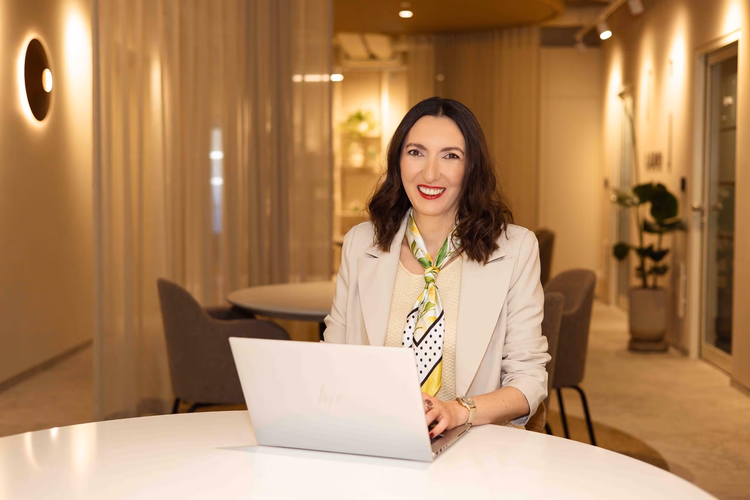 Alicja Dworowska z uśmiechem pracuje na laptopie przy biały stole w nowoczesnym biurze.